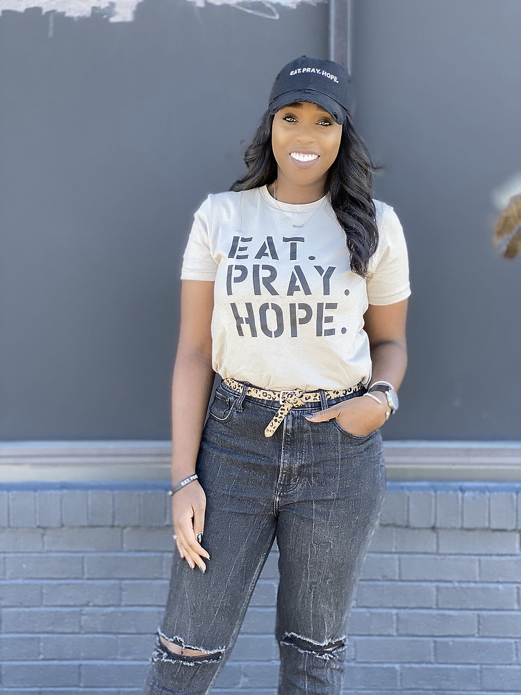 Black woman wearing ‘Eat. Pray. Hope.’ graphic tee and cap standing confidently against a gray wall.